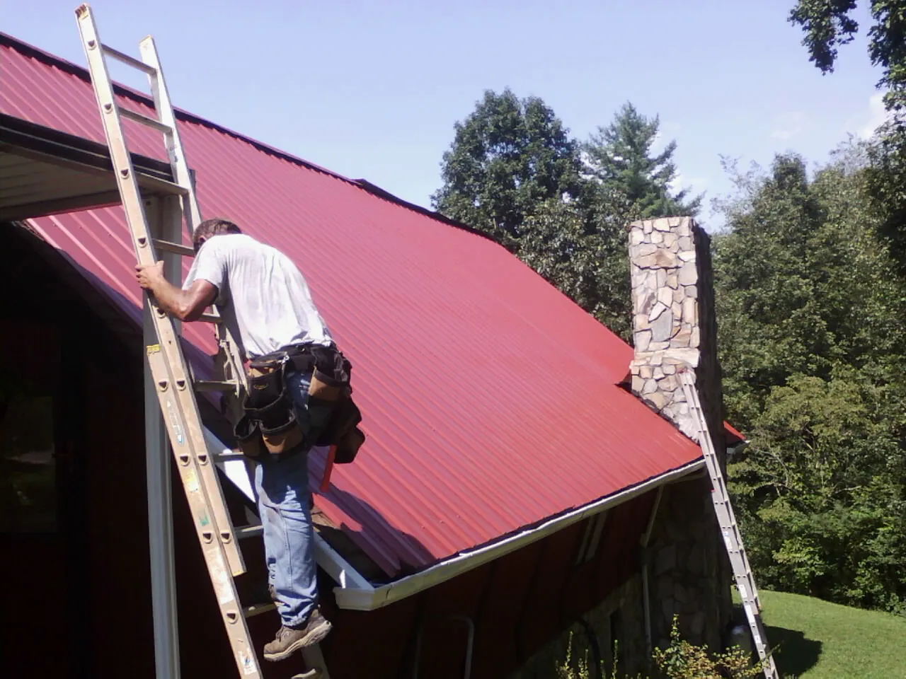 Roofer on a ladder beside a red metal roof and stone chimney — Soffit & Fascia Repair by Roofing Brothers in Glenford
