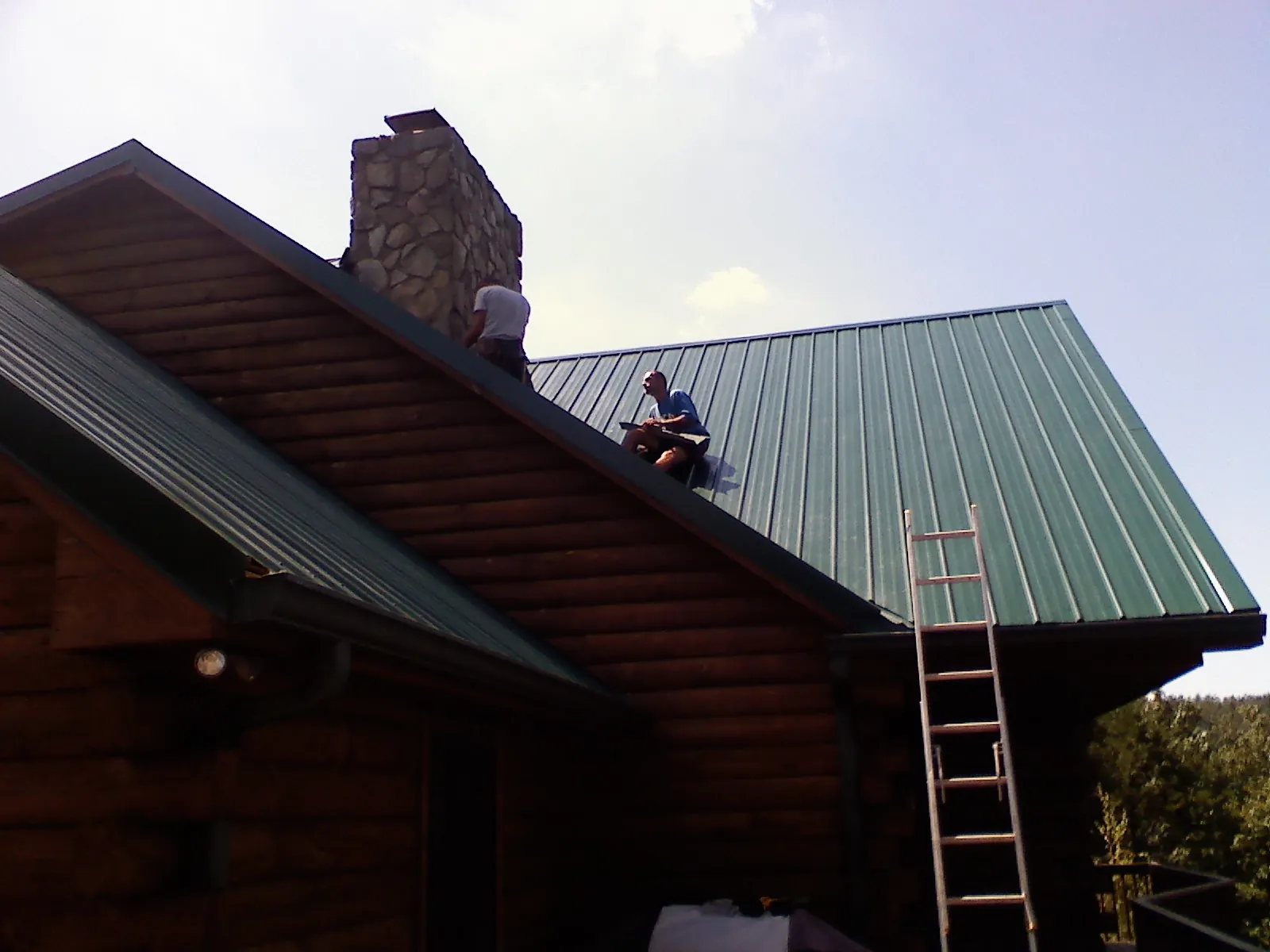 Roofers installing green metal panels beside a stone chimney on a log home in Glenford